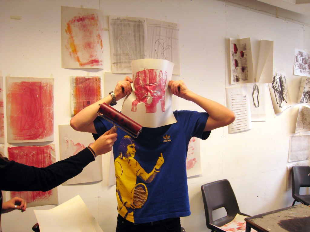 a student holds a sheet of paper over their face, while another runs a roller with red ink over it. workshop at the cass, london met foundation art and design, 2007