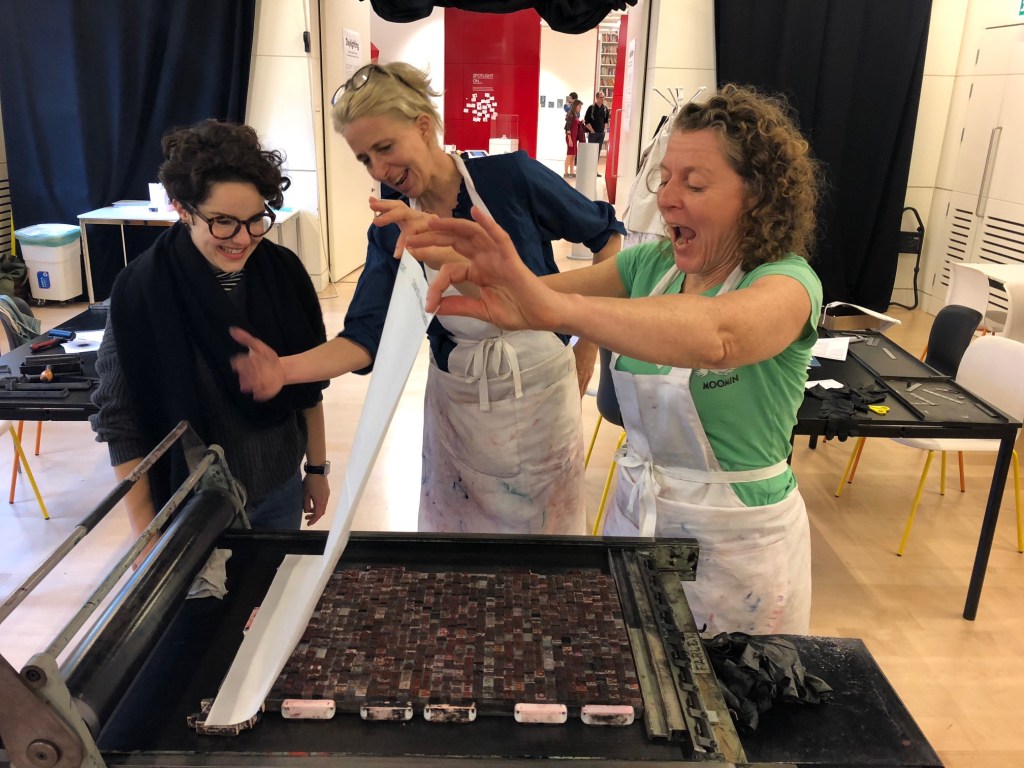 Three women lift a sheet of paper off a printing block of text in a letterpress workshop at the Wellcome Collection.