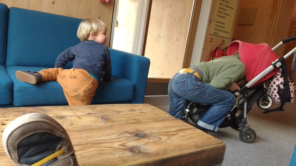 a woman is sticking her head into a red pram, while a toddler climbs on a sofa next to her.