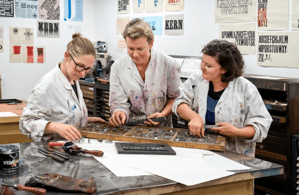 three women stand at a table working with letterpress type. photo David Sandison.