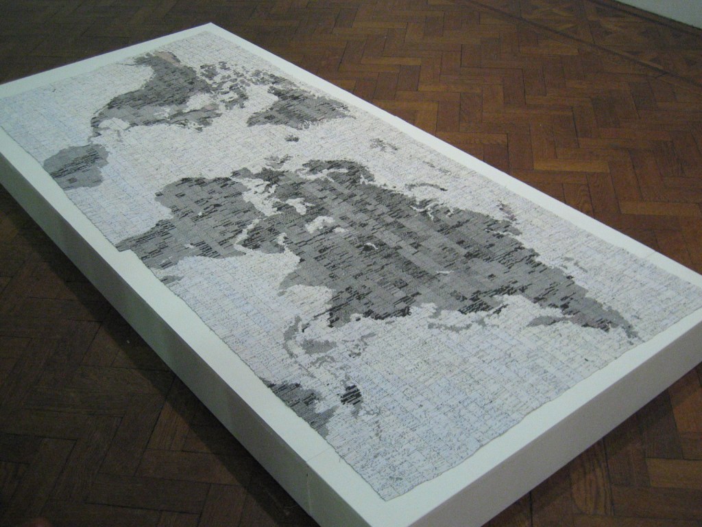 a large incomplete map of the world in black and white on a raised white plinth above a wooden floor.