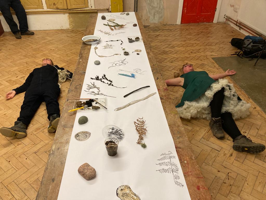 Two women lie on a wooden parquet floor either side of a long table. On the right a woman wearing a bright green dress with a fluffy white underskirt has her arms stretched out wide. On the left a woman in a dark jumpsuit with a tangle of beach rope
