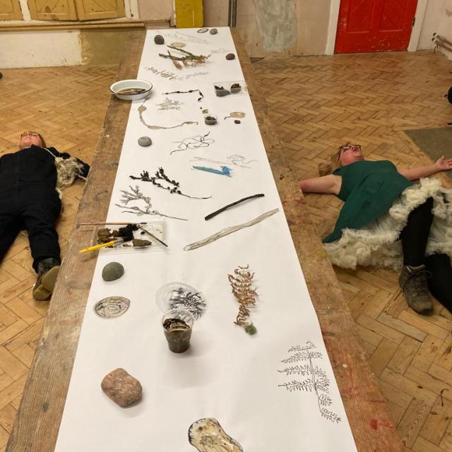 Two women lie on a wooden parquet floor either side of a long table. On the right a woman wearing a bright green dress with a fluffy white underskirt has her arms stretched out wide. On the left a woman in a dark jumpsuit with a tangle of beach rope. The table in the centre is covered with beach finds like seaweed stones and driftwood, each object has a drawing of itself next to it.