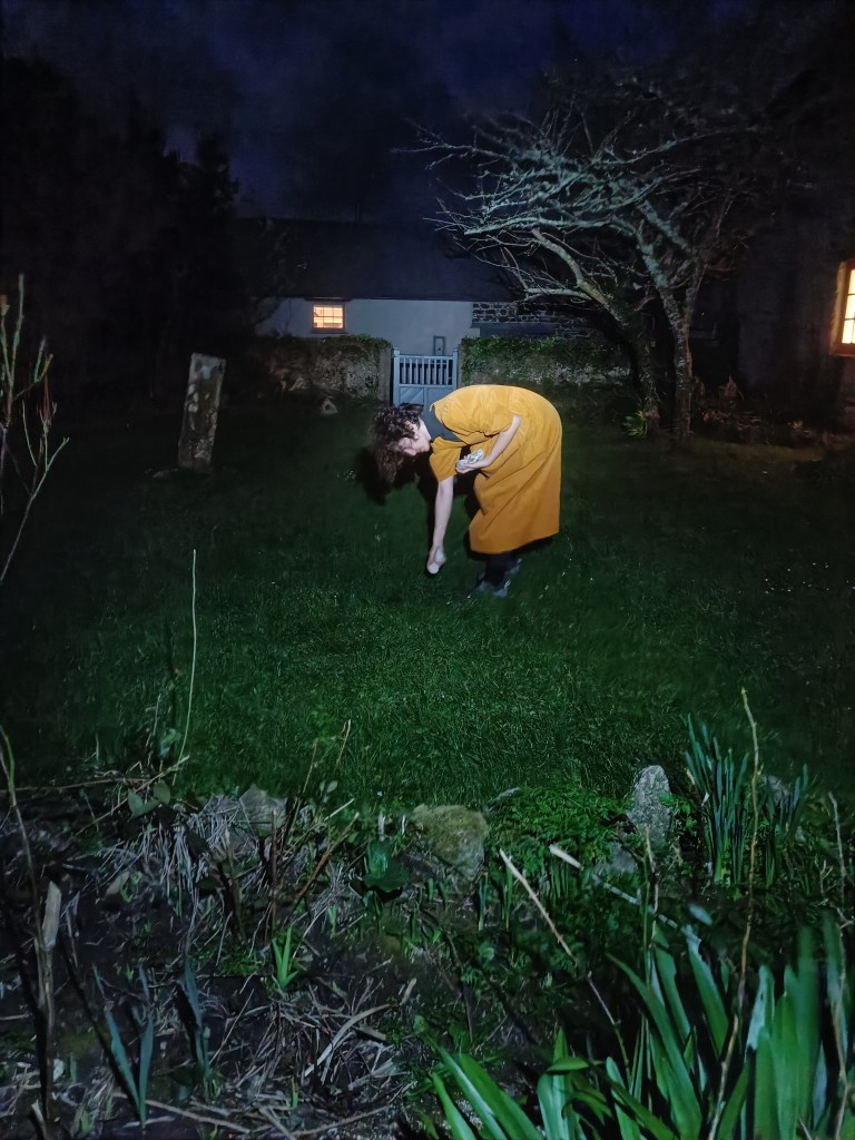 a woman in a yellow dress in a dark garden. she is brightly lit. she is bending over to place a white stone, and is holding more stones in her arms. in the background a single window is illuminated yellow.