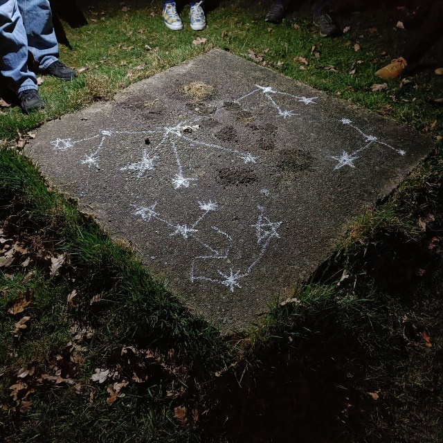 a square of paving surrounded by grass with chalk stars drawn on it, connected by lines into constellations. The feet of people standing around the square are visible